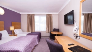 Cosy hotel room with twin beds, purple accents, flat-screen TV, desk, and chair. Warm lighting creates a welcoming atmosphere.