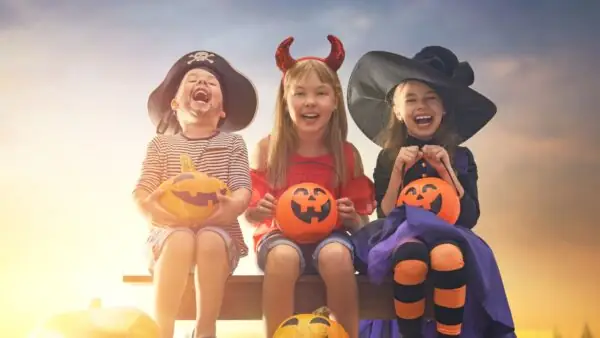 Three children laugh in Halloween costumes, sitting with pumpkin lanterns on a wooden bench against a sunset sky.