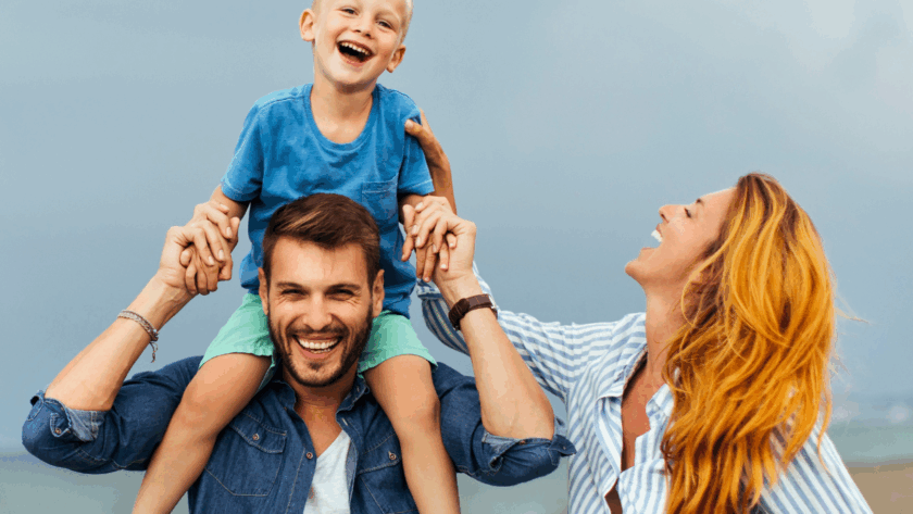 A smiling man carries a laughing boy on his shoulders while a woman stands beside them on a beach, all appearing happy.
