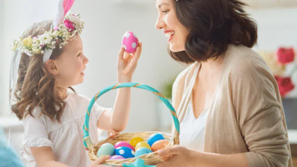 A woman and a girl in flower crowns hold a basket of colorful Easter eggs. As the girl, wearing bunny ears, shows off a pink egg, they chat excitedly about finding the best hotel deals for their spring holiday.