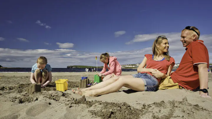 Family enjoys sunny beach day, children build sandcastles, parents relax on sandy shore, blue sky backdrop.