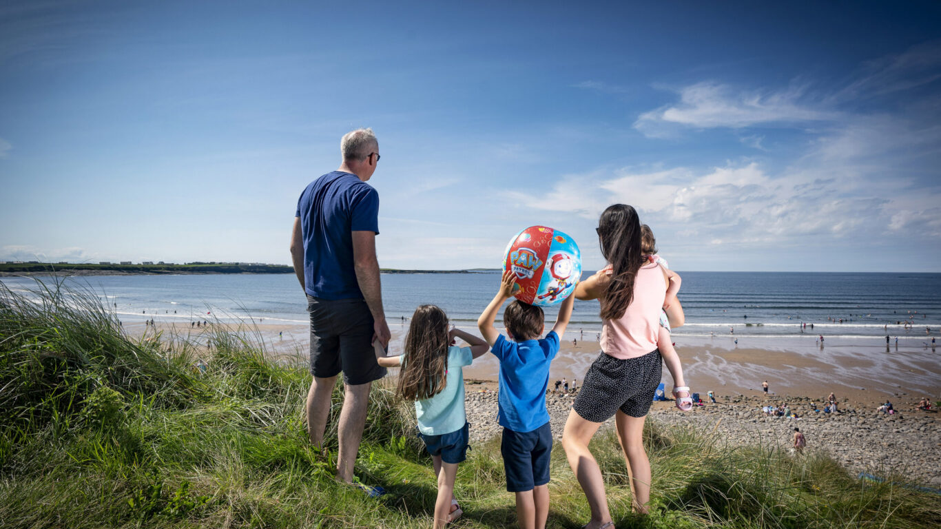 Family enjoys scenic beach view, grassy dunes, and lively shoreline, perfect for a seaside holiday at our welcoming hotel.